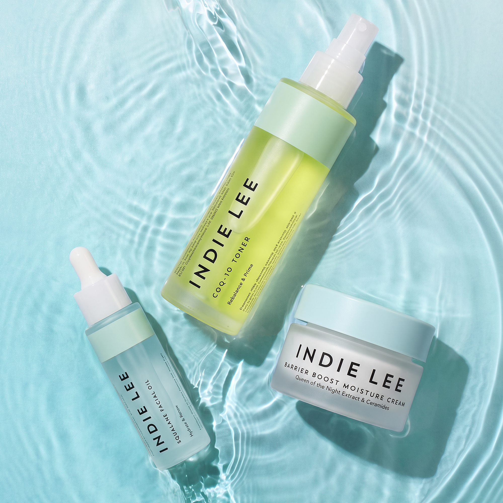Three Indie Lee skincare products arranged on a light aqua water surface.