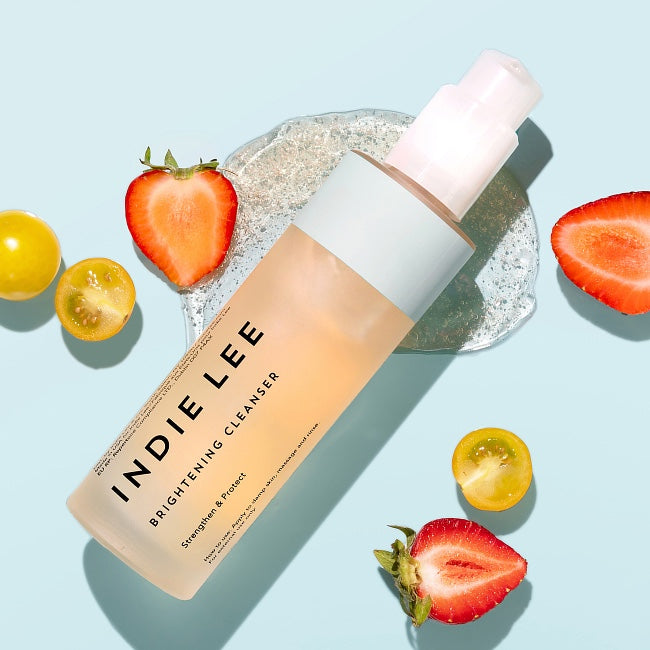 Indie Lee Brightening Cleanser bottle with strawberries on a light blue background