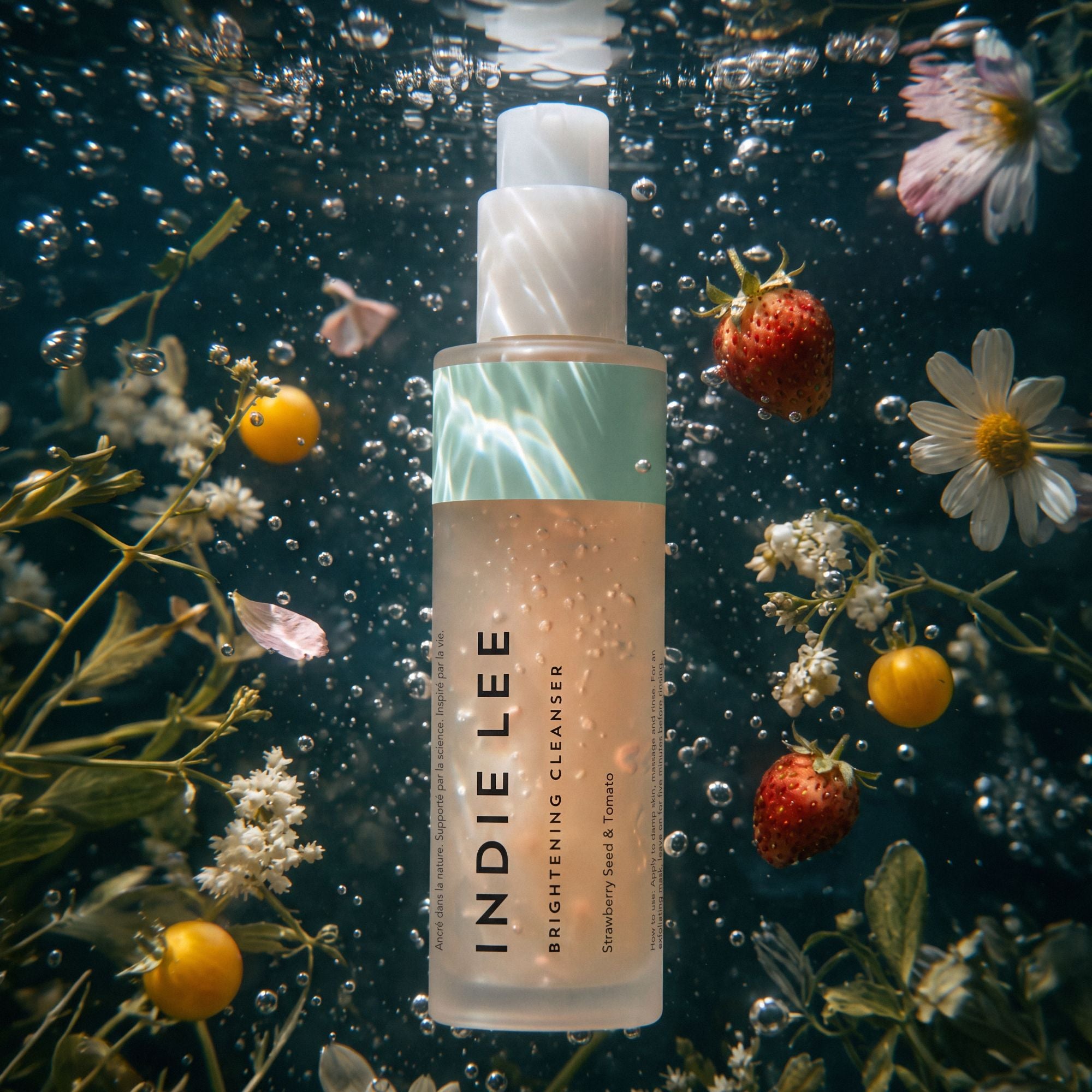 A bottle of Indie Lee Brightening Cleanser floating underwater in clear water with bubbles, surrounded by strawberries, white daisies, and yellow flowers.