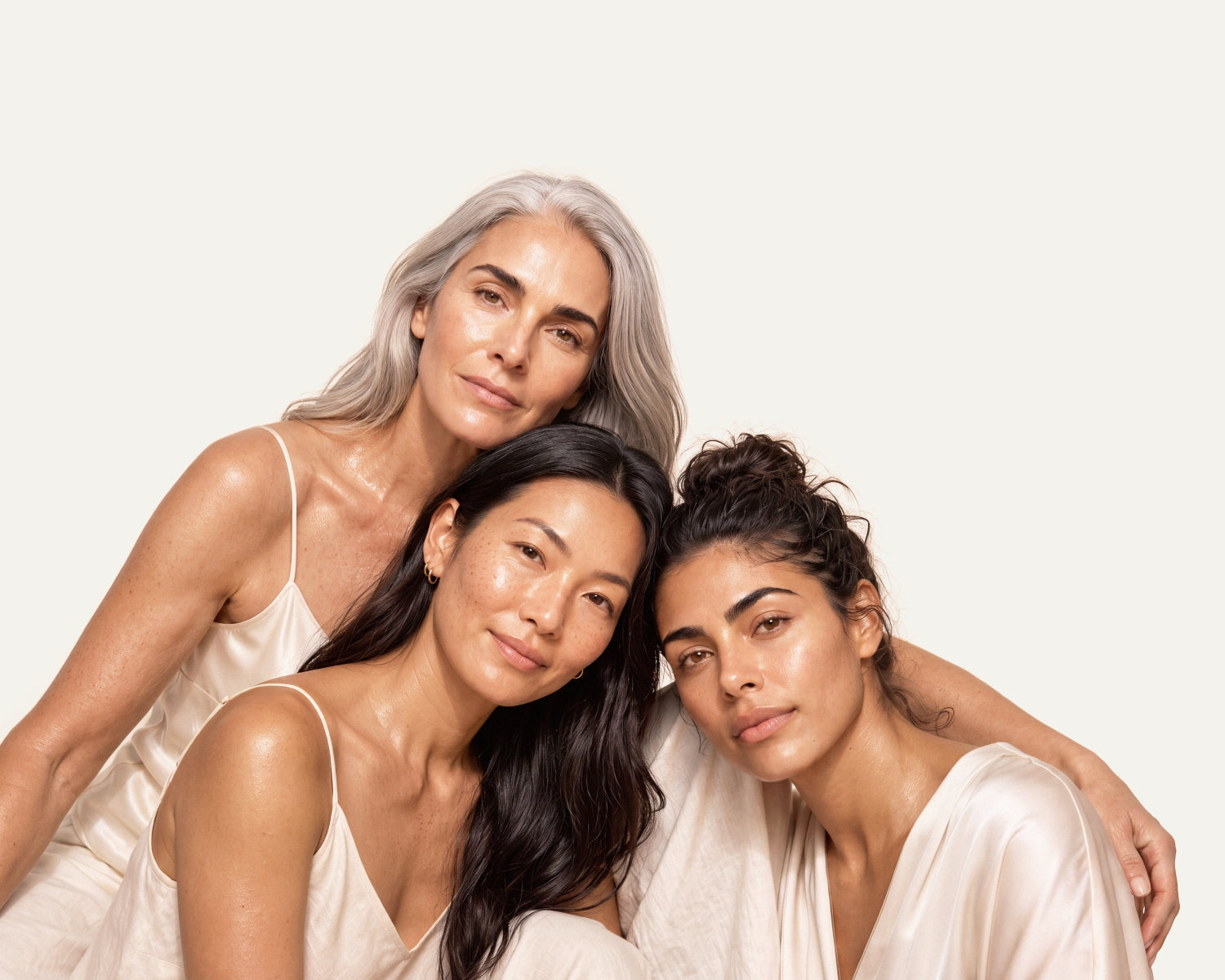 Three women with natural skin tones and varied hair colors (gray, black, and brown) wearing matching pale cream satin camisoles, posed closely together against a solid off-white background.