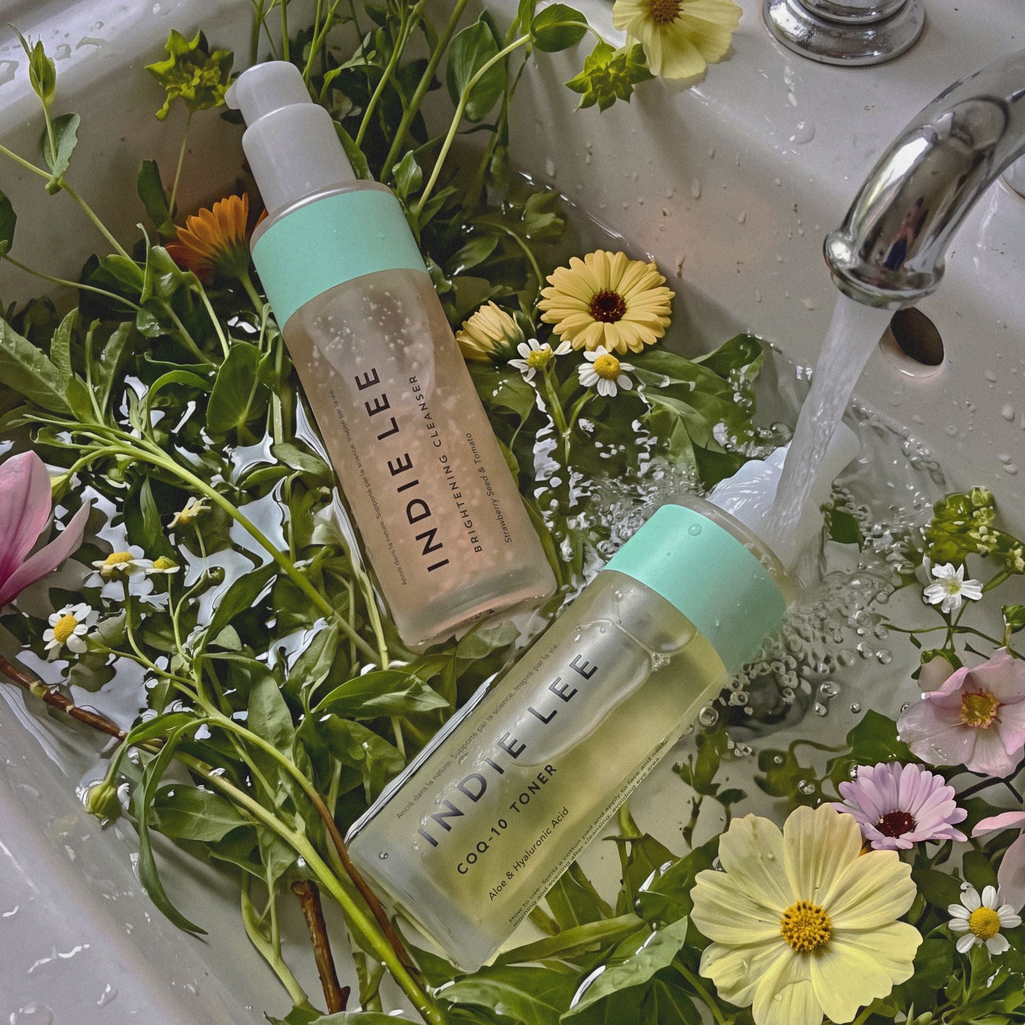 Two Indie Lee skincare bottles placed in a sink filled with water, green leaves, and colorful wildflowers.