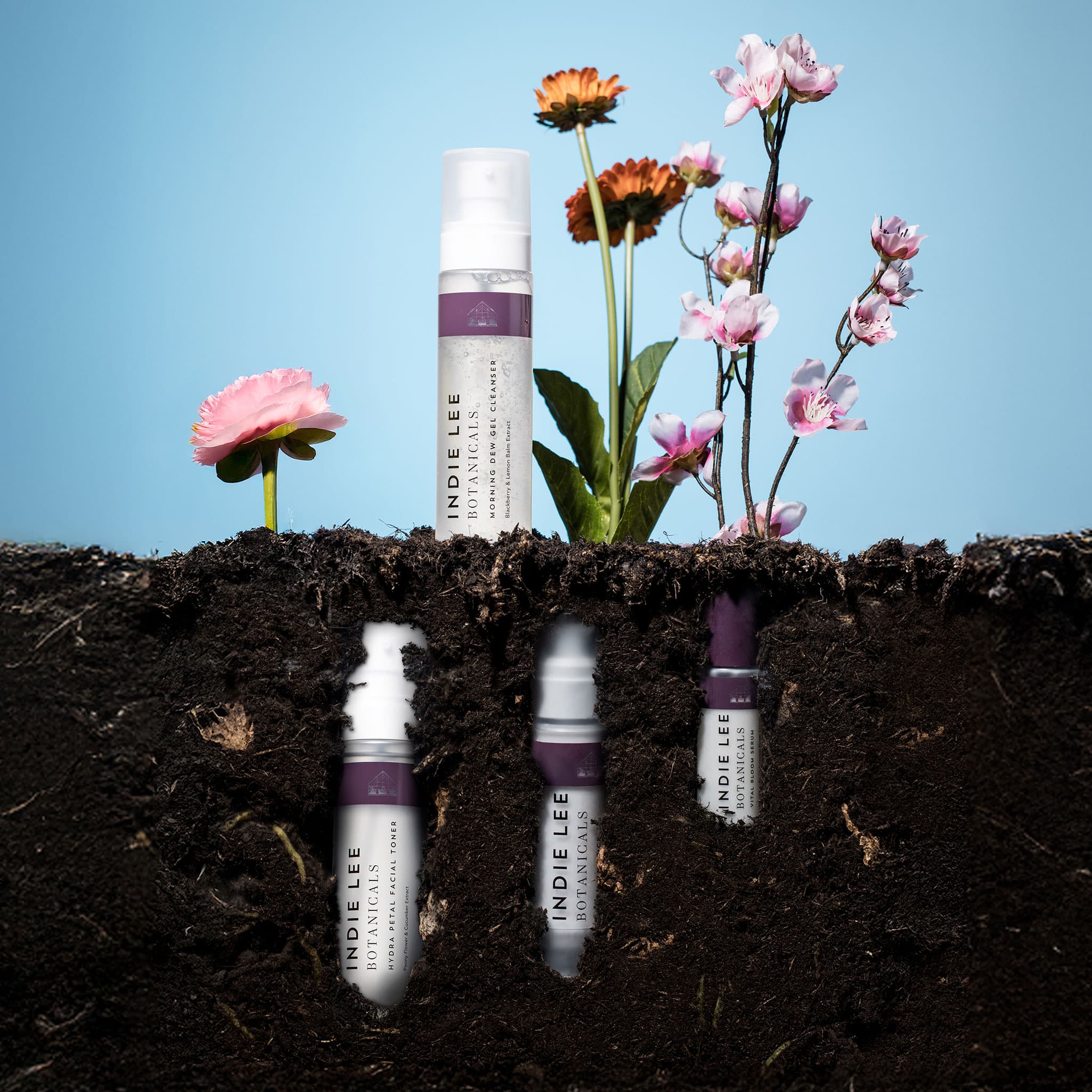 Indie Lee skincare products with flowers emerging from soil against a blue sky