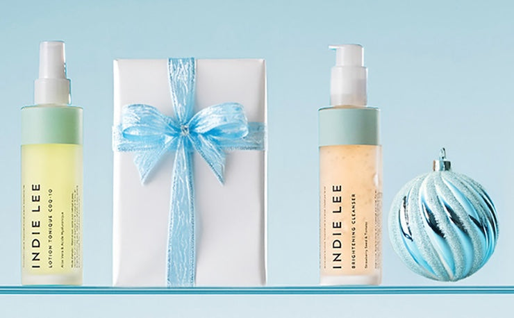 Two bottles of Indie Lee skincare products with a wrapped gift and ornament on a light blue background