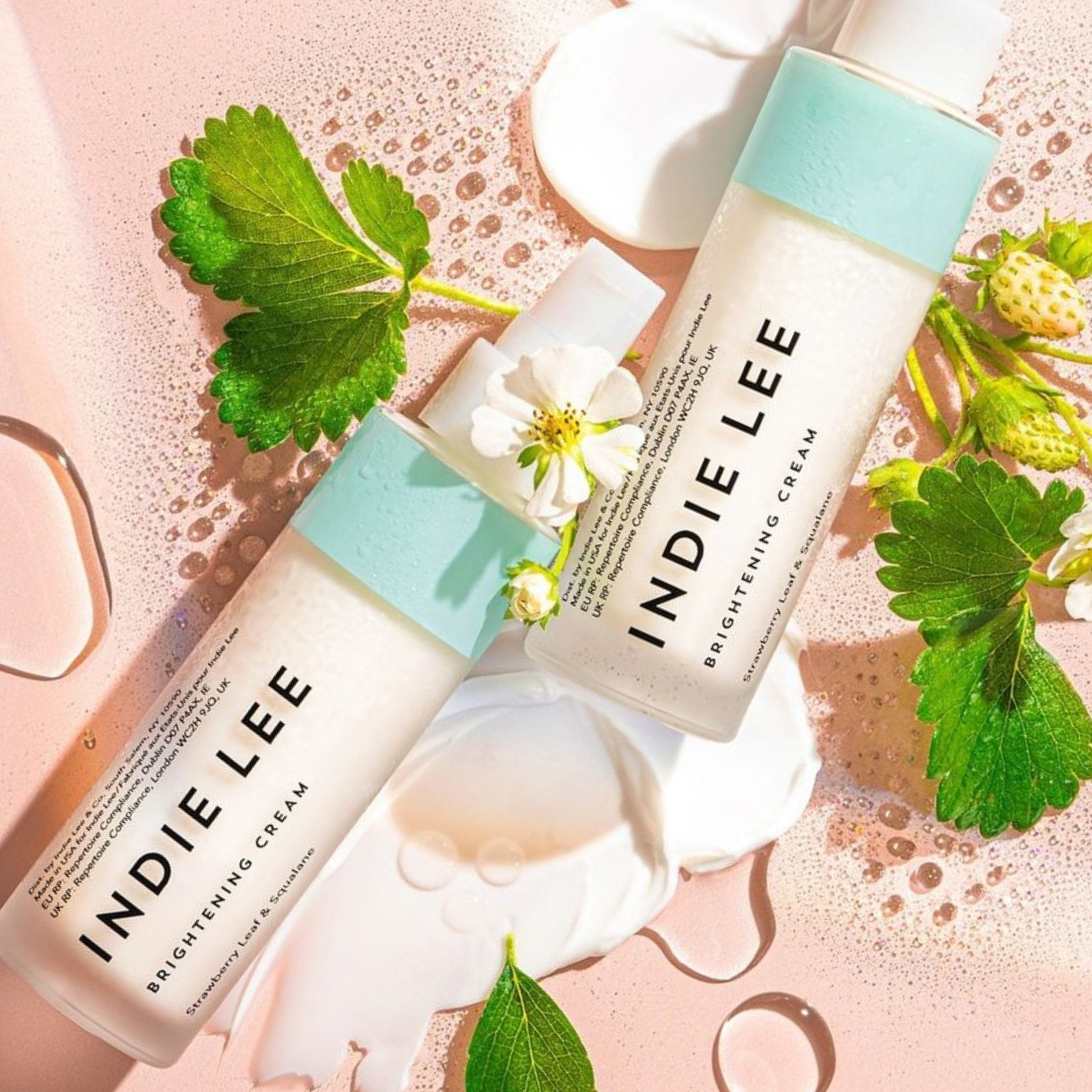 Two bottles of Indie Lee Brightening Cream with green leaves and flowers on a pink background