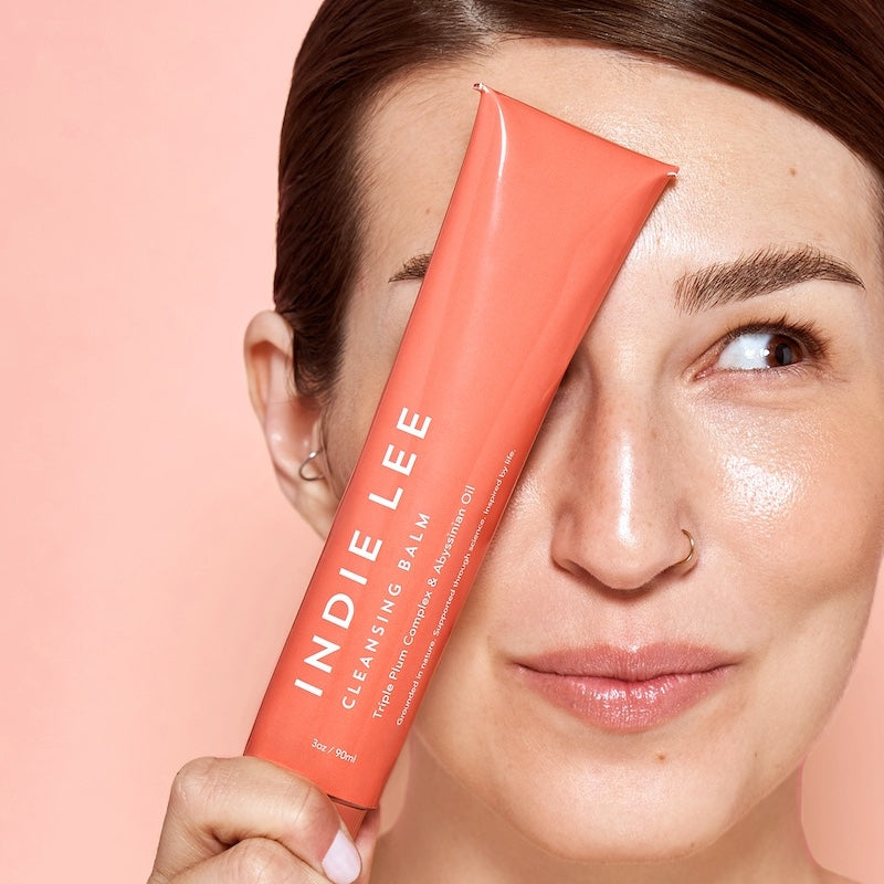 Woman holding a pink tube of Indie Lee Cleansing Balm next to her face