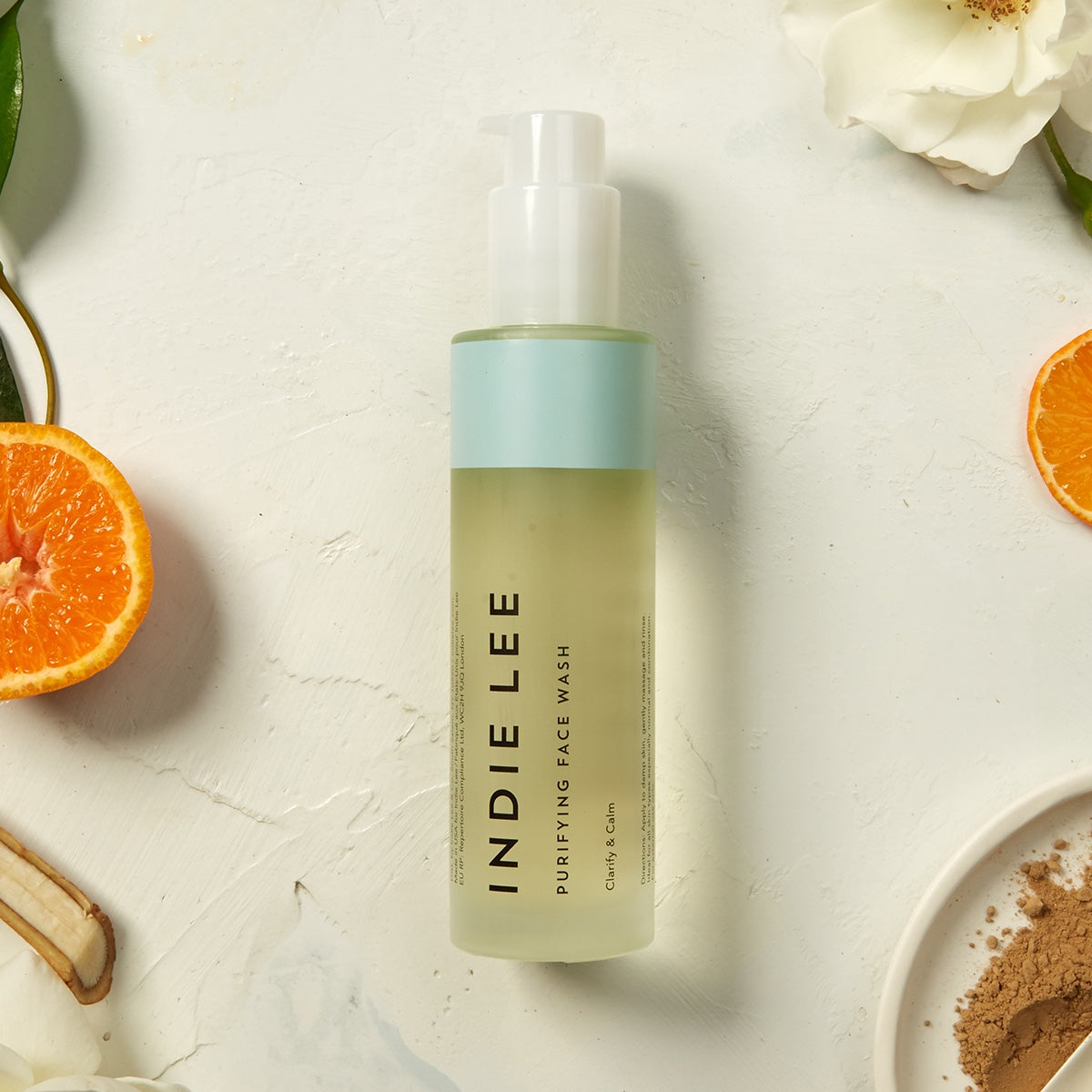 Indie Lee face Purifying Face Wash bottle on a light background with oranges and flowers