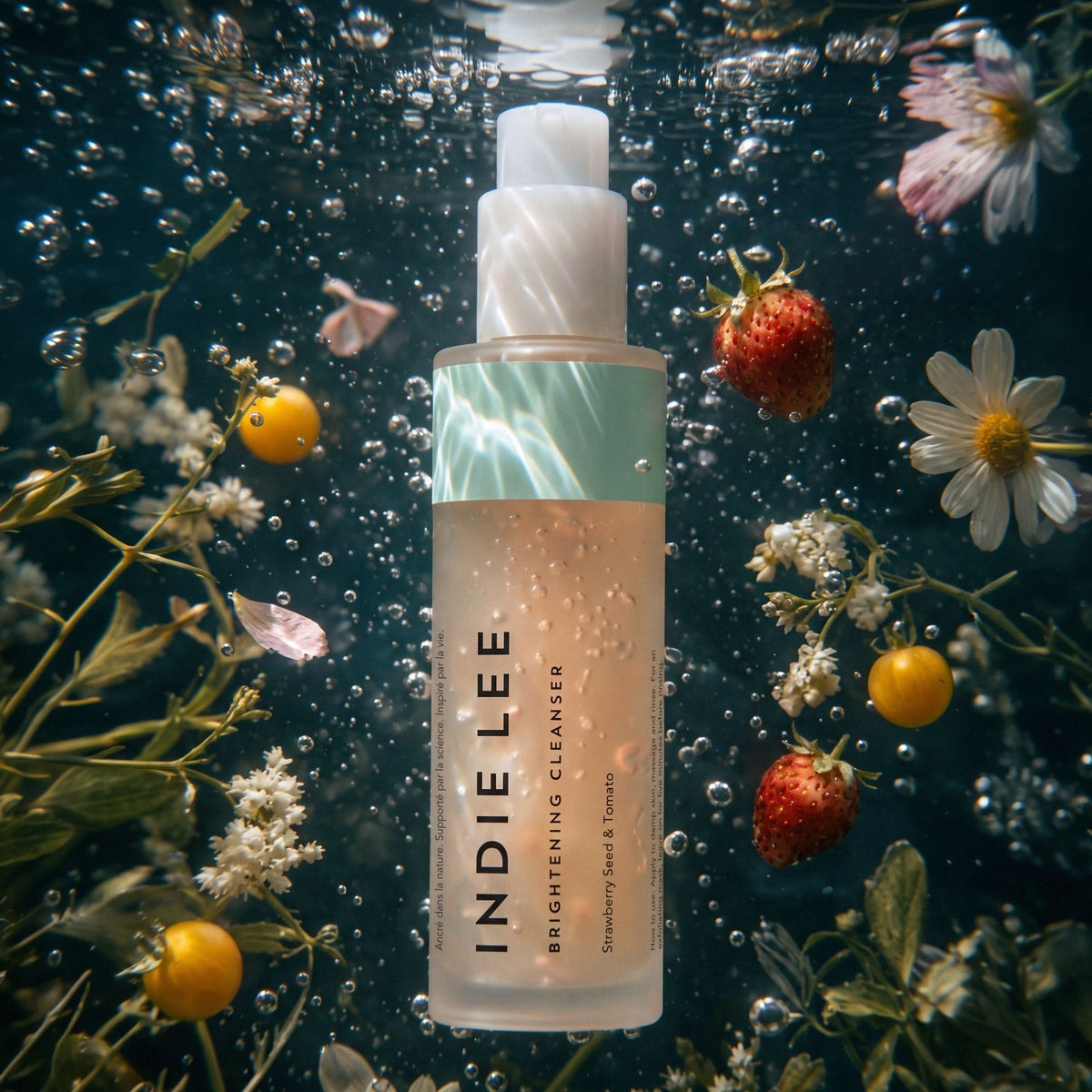 A bottle of Indie Lee Brightening Cleanser floating underwater in clear water with bubbles, surrounded by strawberries, white daisies, and yellow flowers.