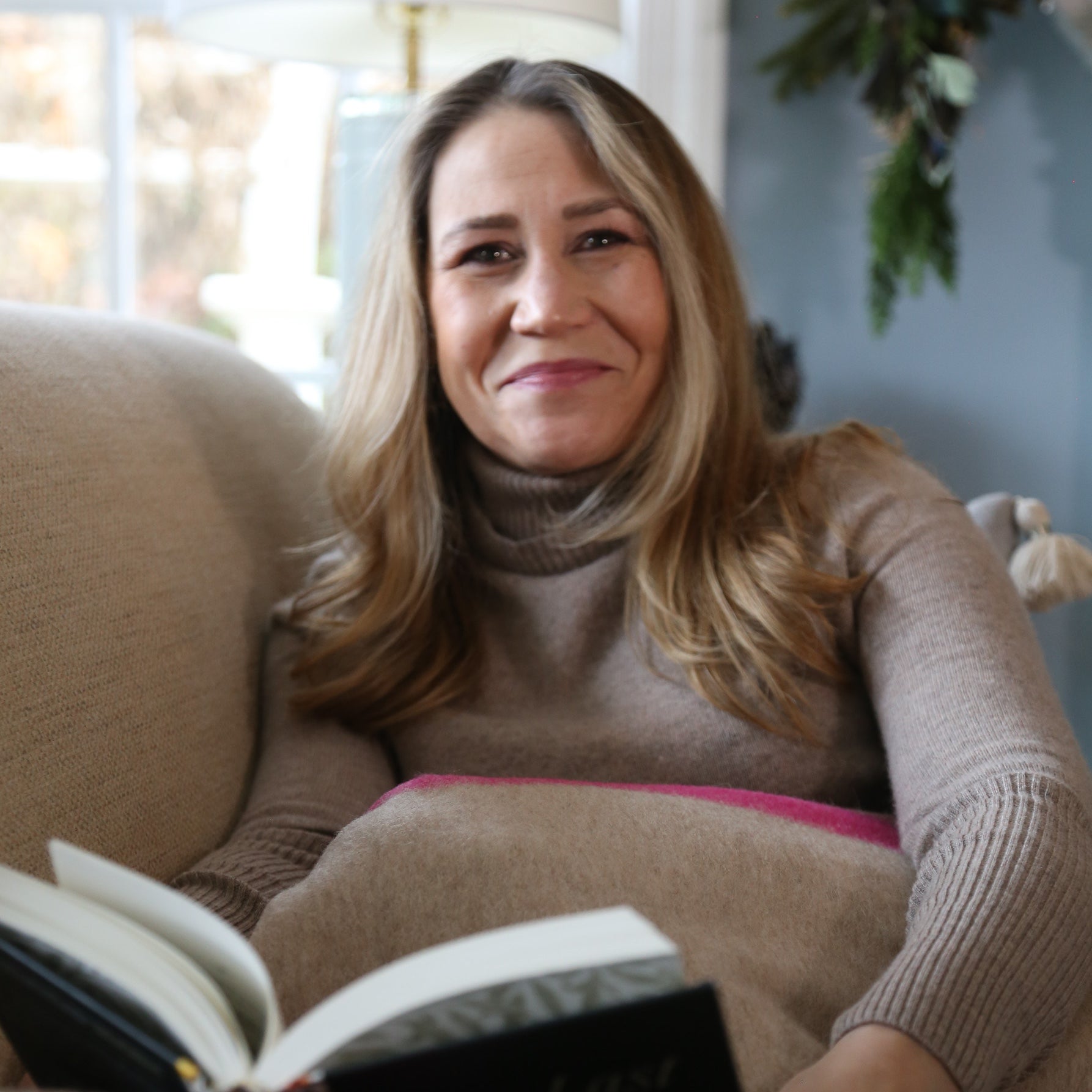Indie Lee sitting on a couch reading a book in a cozy living room